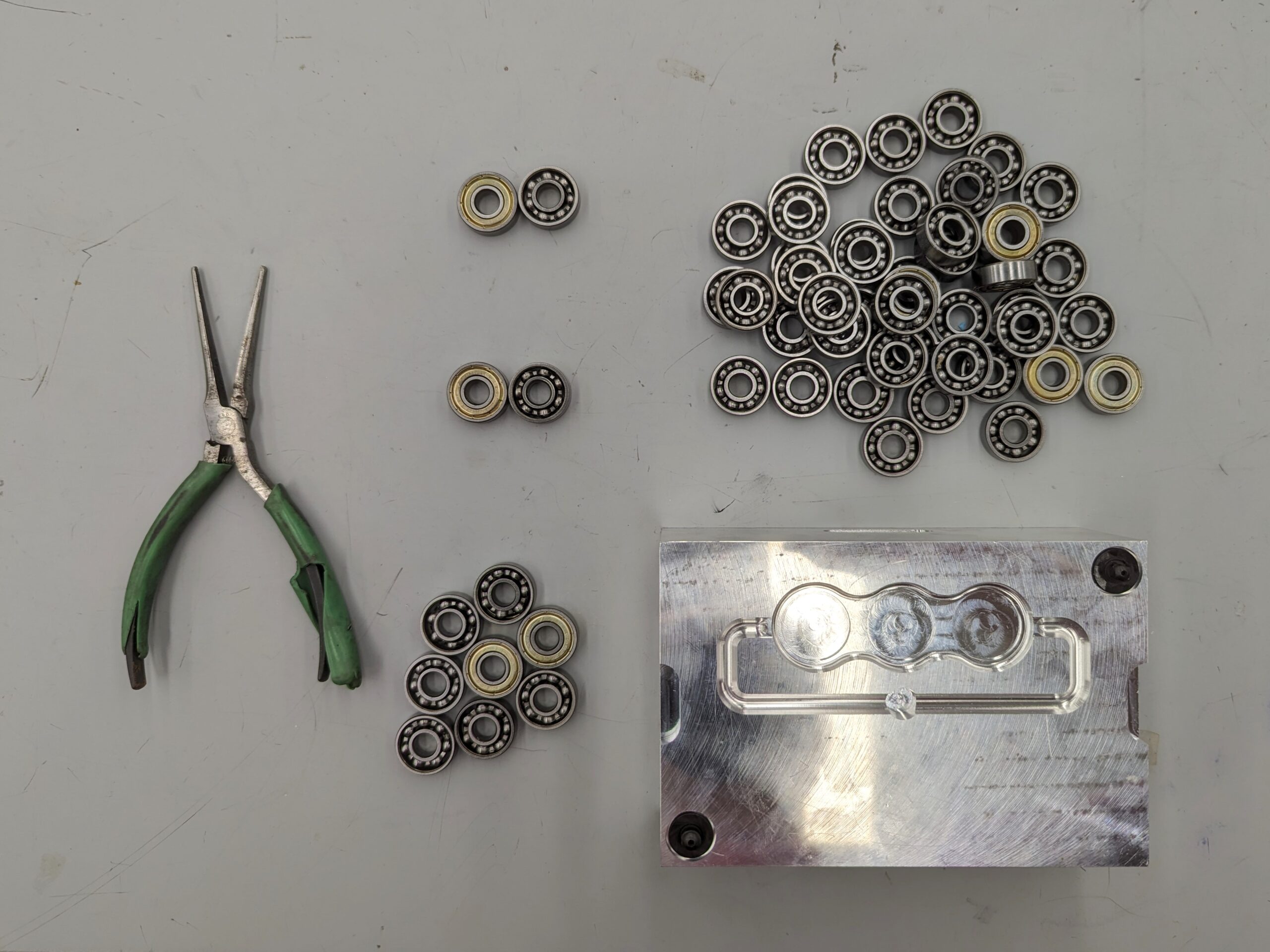 Pliers, 2 sets of bearings, a small pile of bearings, and a large pile of bearings sitting by an aluminum block mold to show that many bearings do not fit into the mold