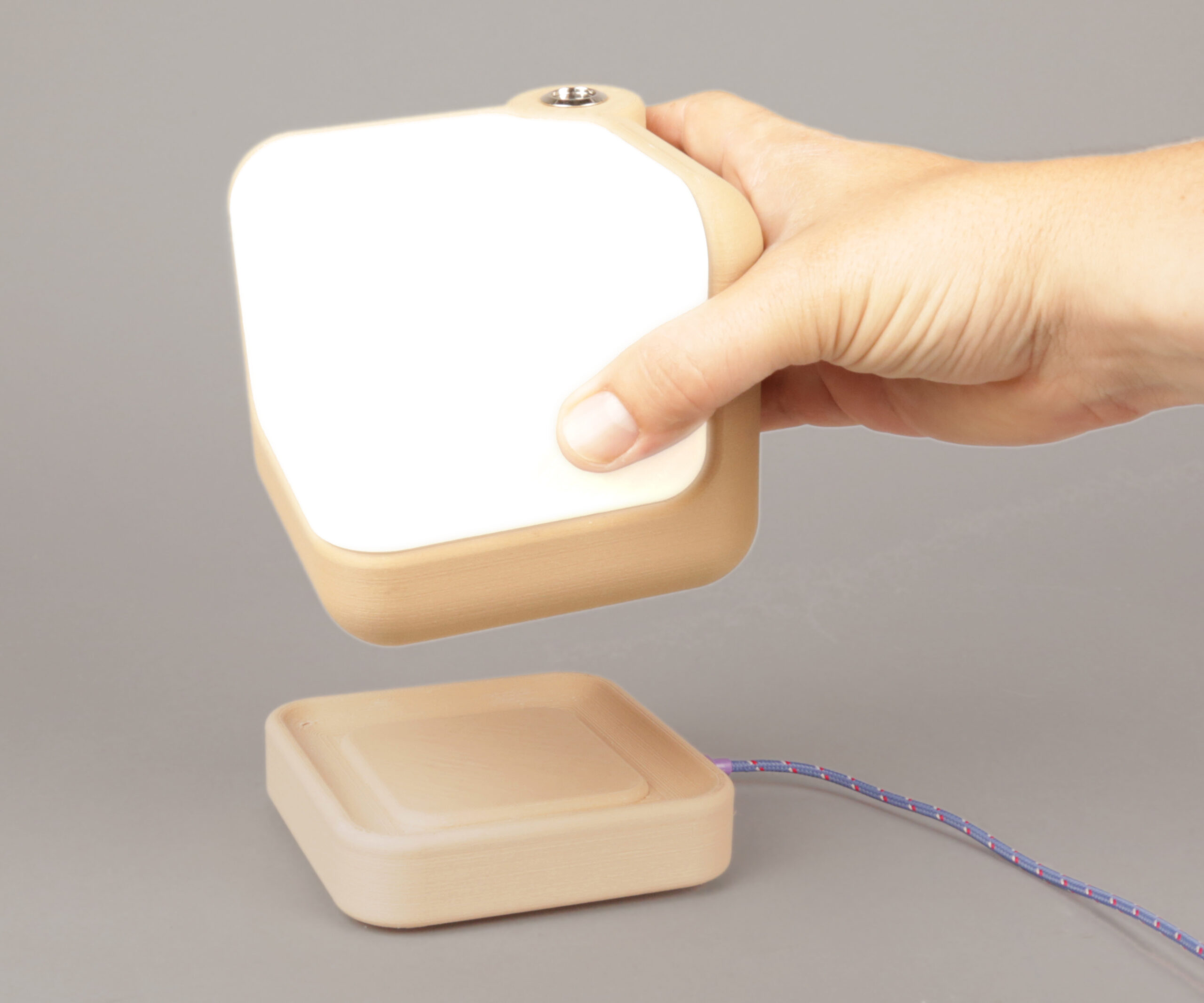 Hand holding cube lamp with rounded edges above a square wireless charging base with rounded edges