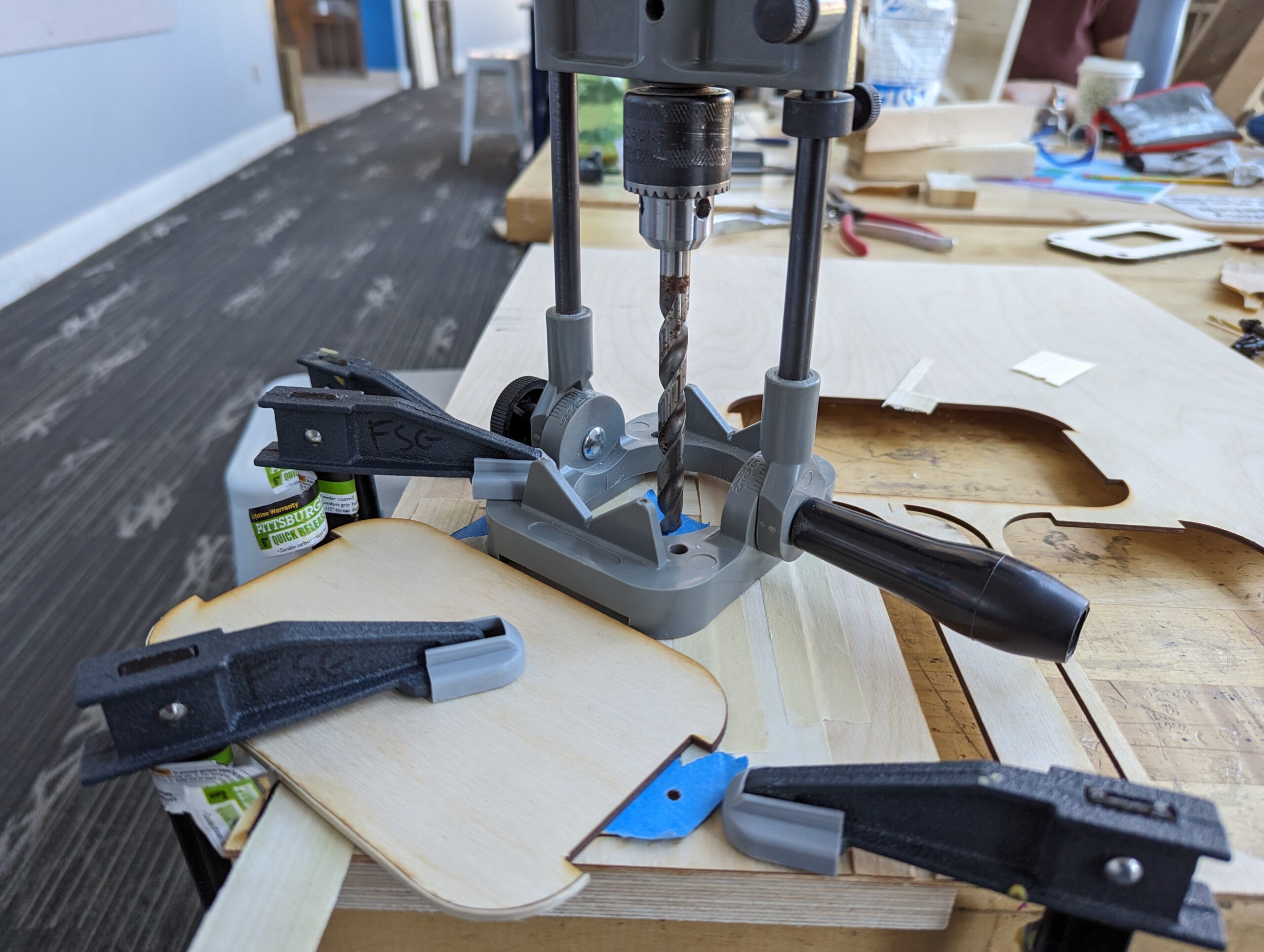 Messy workbench with clamps holding wood piece down and a drill jig centering a ground-down drill bit to add countersinks to holes