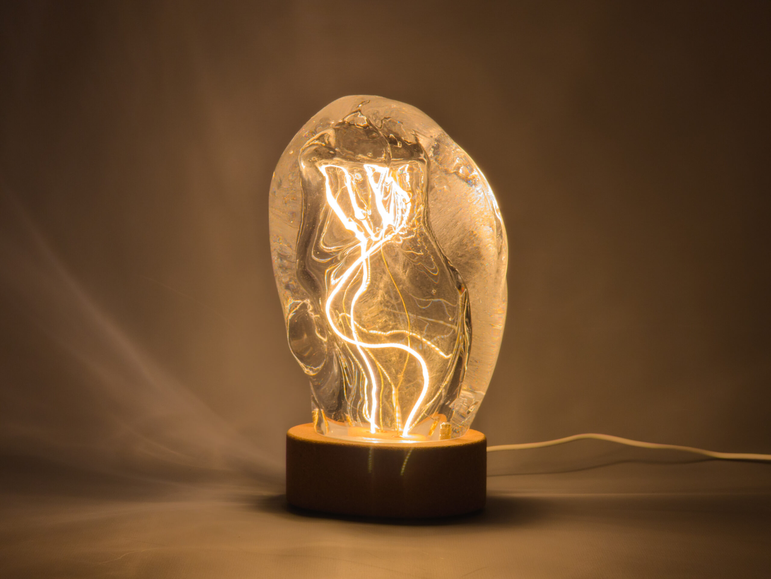 Organically-shaped blown glass lamp with obround cork base and curled LED filament lighting the glass from inside.