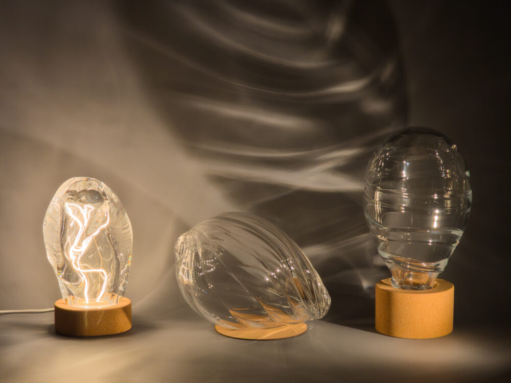 Three transparent glass forms with cork bases set beside each other against a gray backdrop. Light shining through the glass projects patterns on the backdrop.