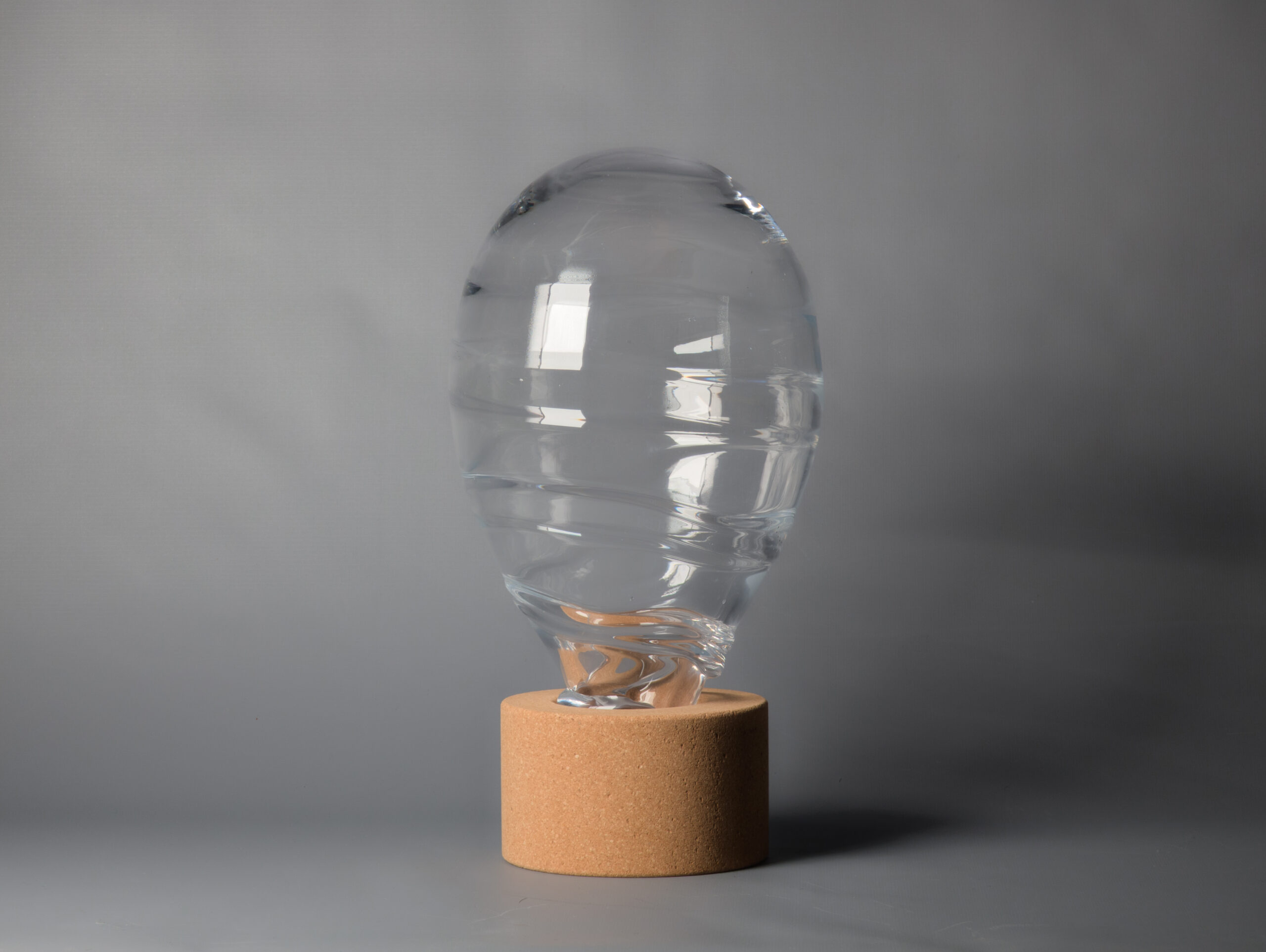 Transparent blown glass bulb with rippled surface and cylindrical cork base set against gray backdrop.