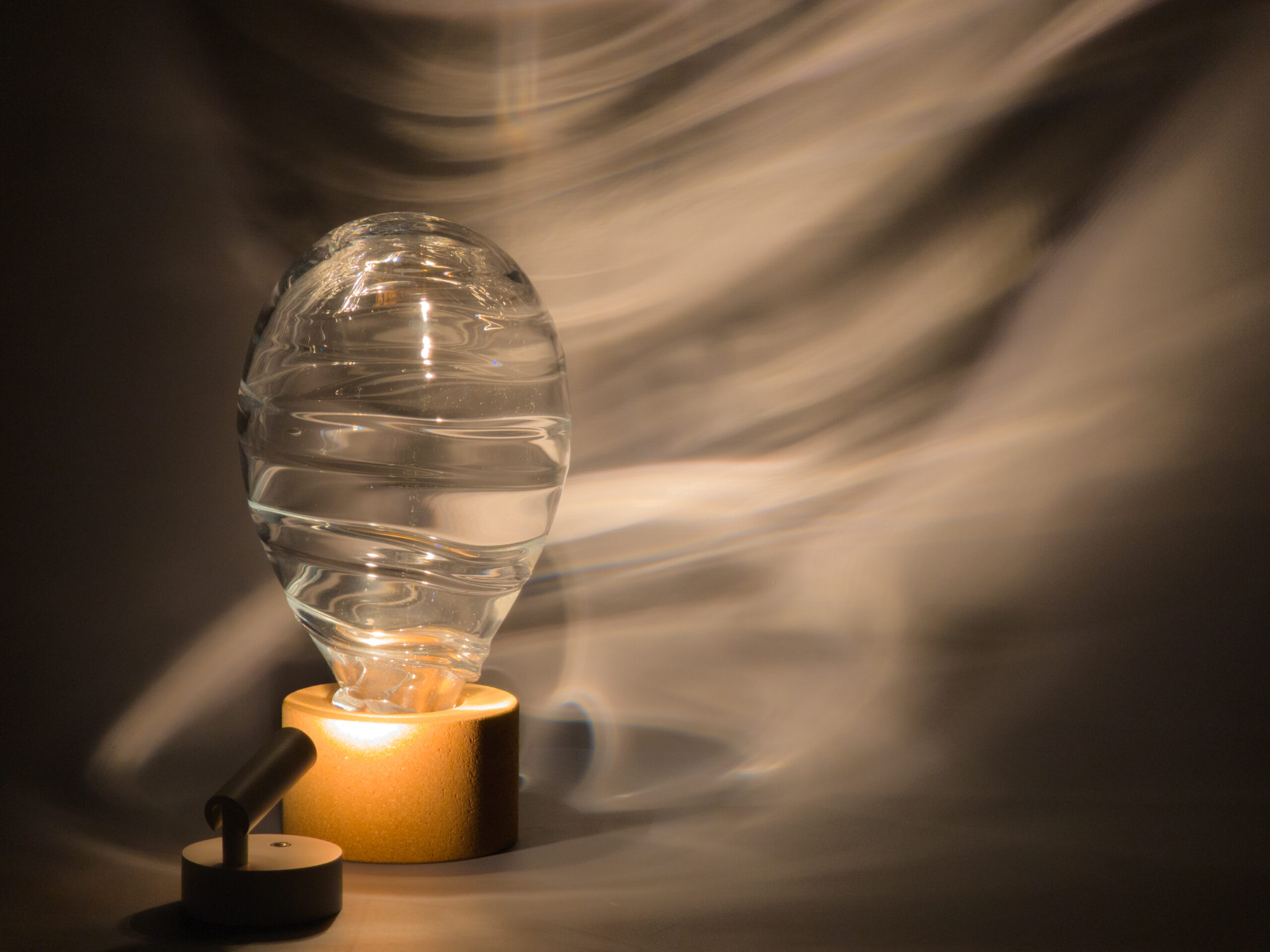 Light shining through a transparent, balloon-like, blown glass and cork sculpture, projecting rippling light onto a gray backdrop.