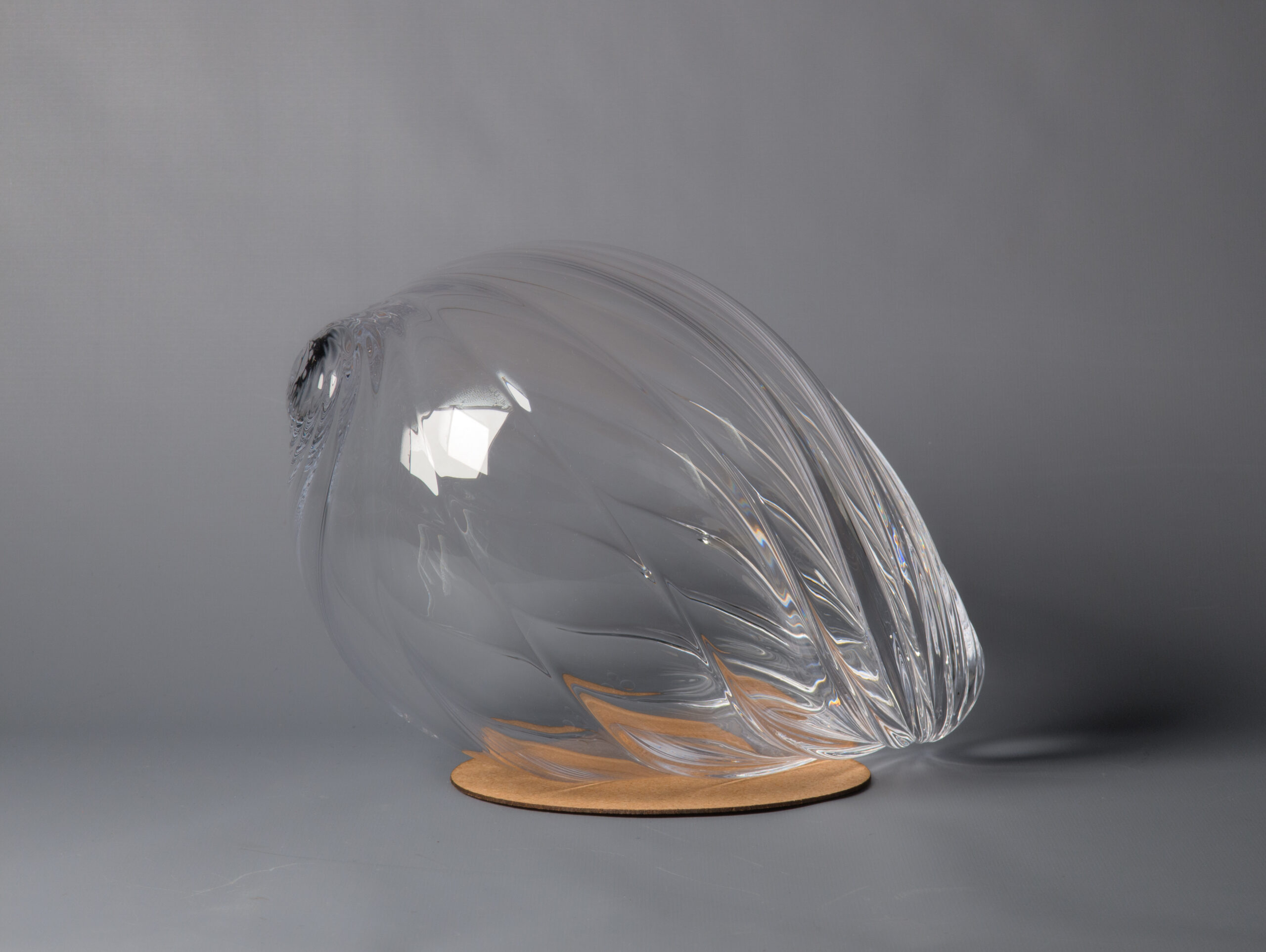 Blown glass bulb with twisted ridges resting on a circular cork sheet against a gray backdrop.