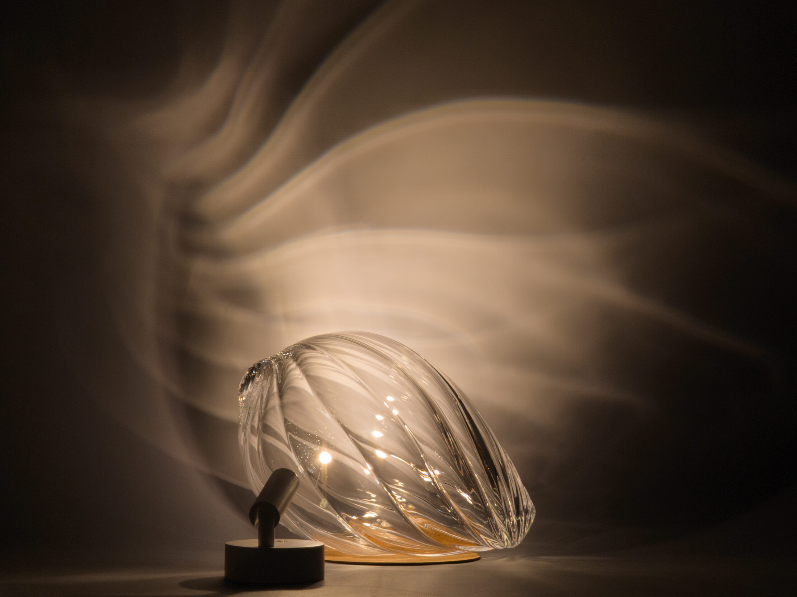 Light shining through a glass form with spiraling fluted texture, projecting a regular pattern of curves onto a gray backdrop.