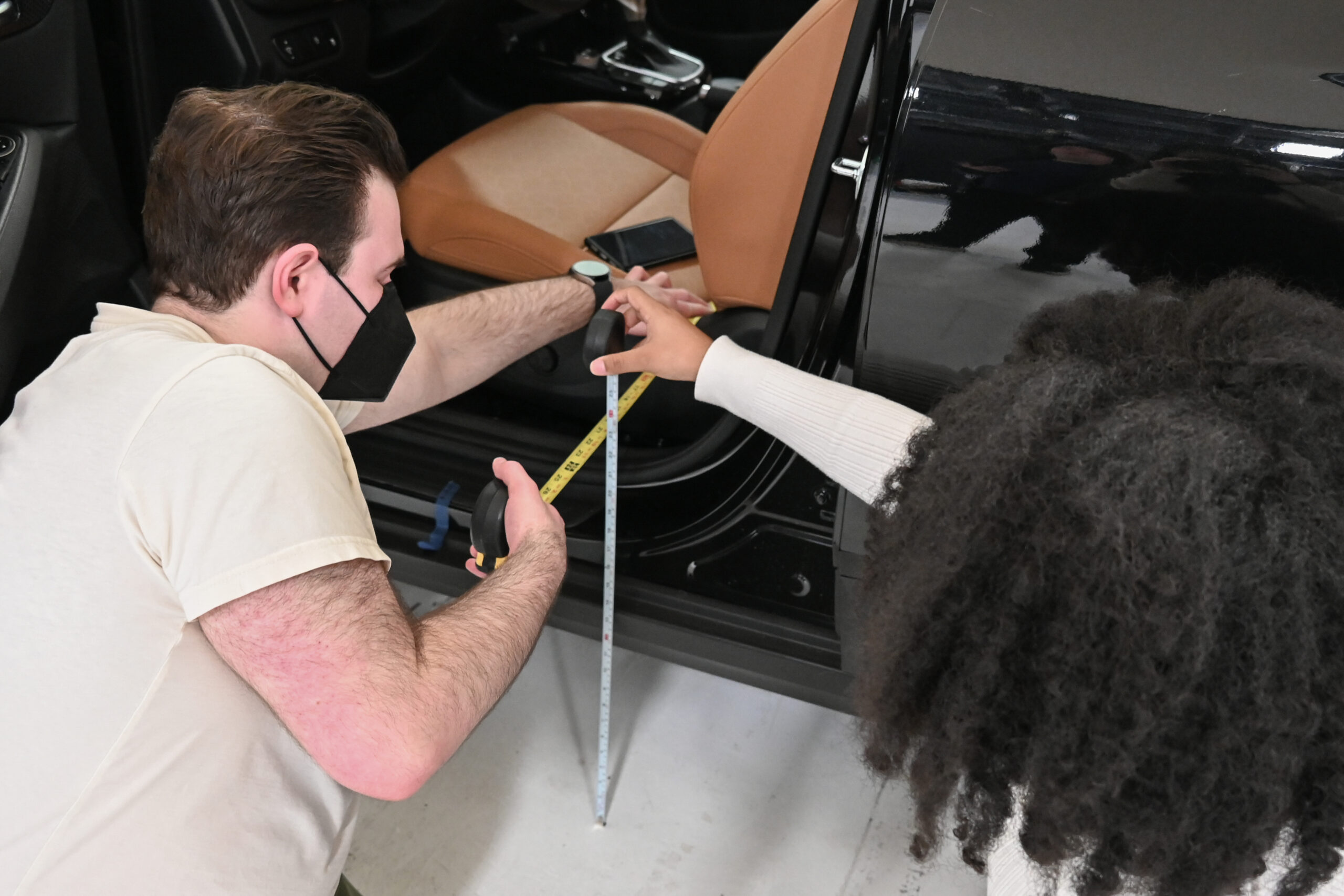 Two people holding measuring tapes against the seat and side of a black car.