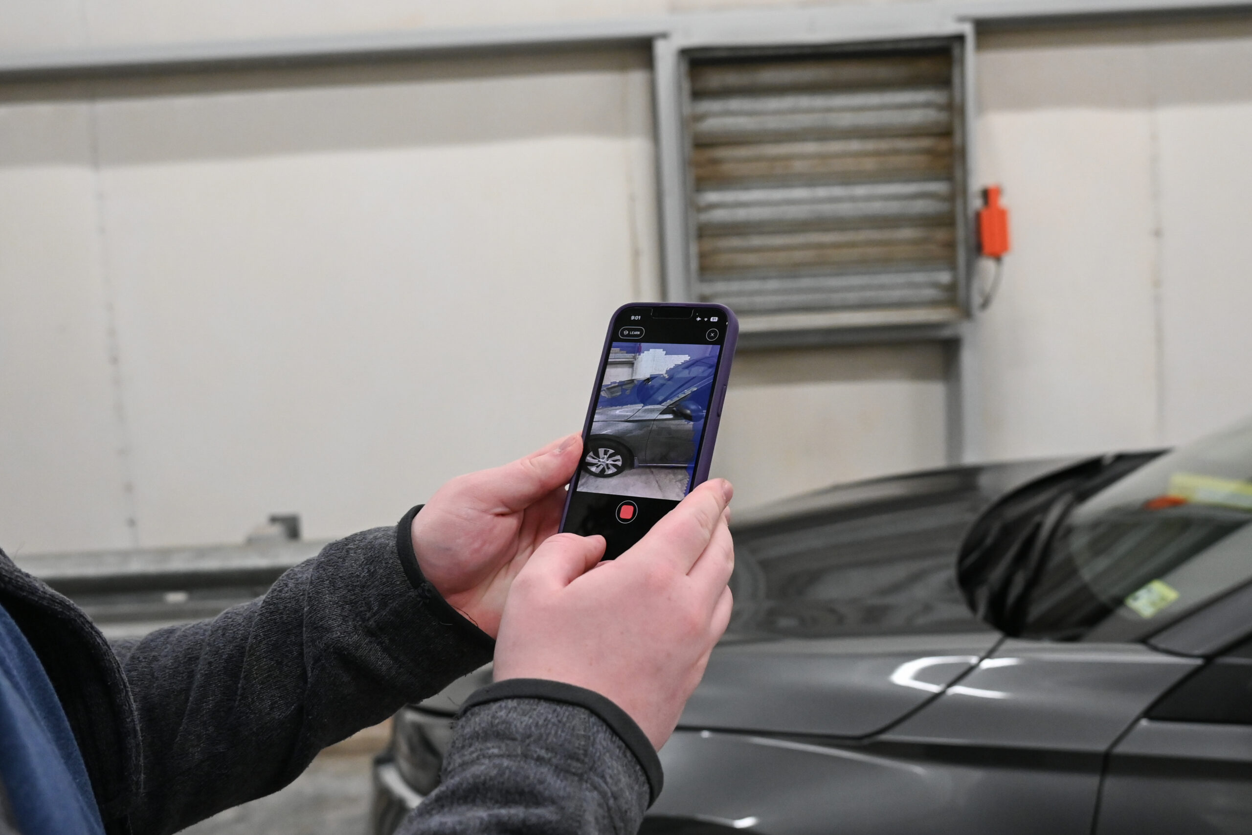 Hands holding a smartphone in front of a gray car. The phone screen shows the phone's camera view while taking a 3d scan of the car.